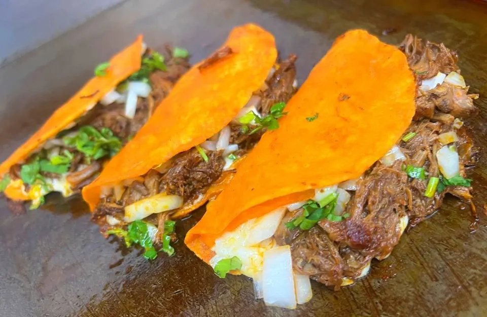 Three birria tacos with red-dipped tortillas, shredded beef, onions, and cilantro on the griddle.