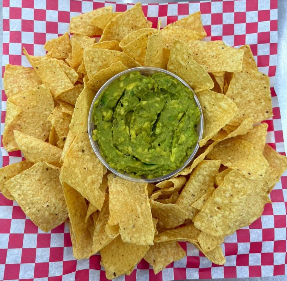 A cup of chunky guacamole surrounded by tortilla chips on red and white checkered paper.