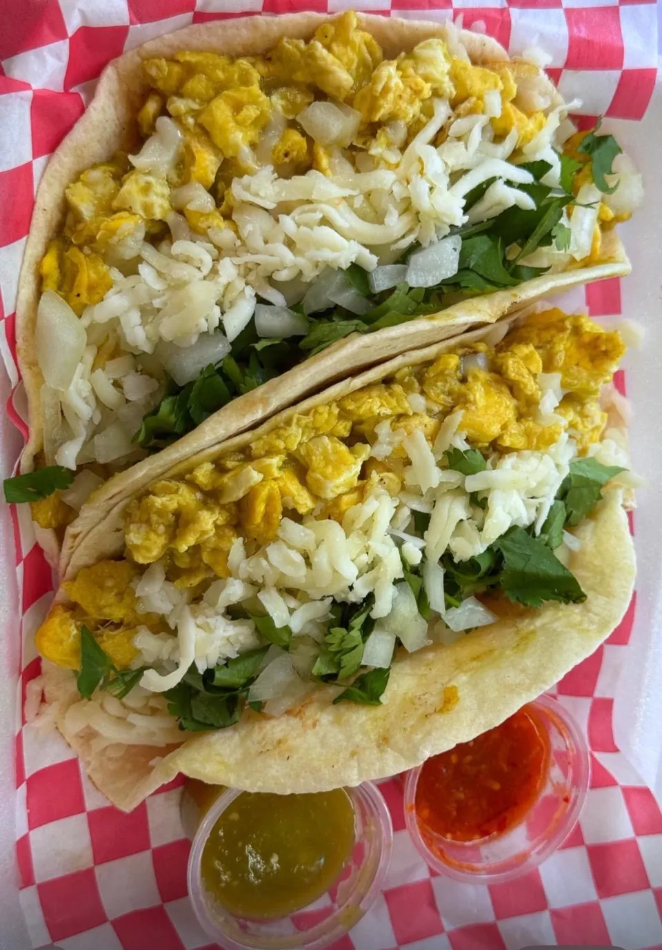 Two breakfast tacos with scrambled eggs, cheese, and cilantro, served with red and green salsa.