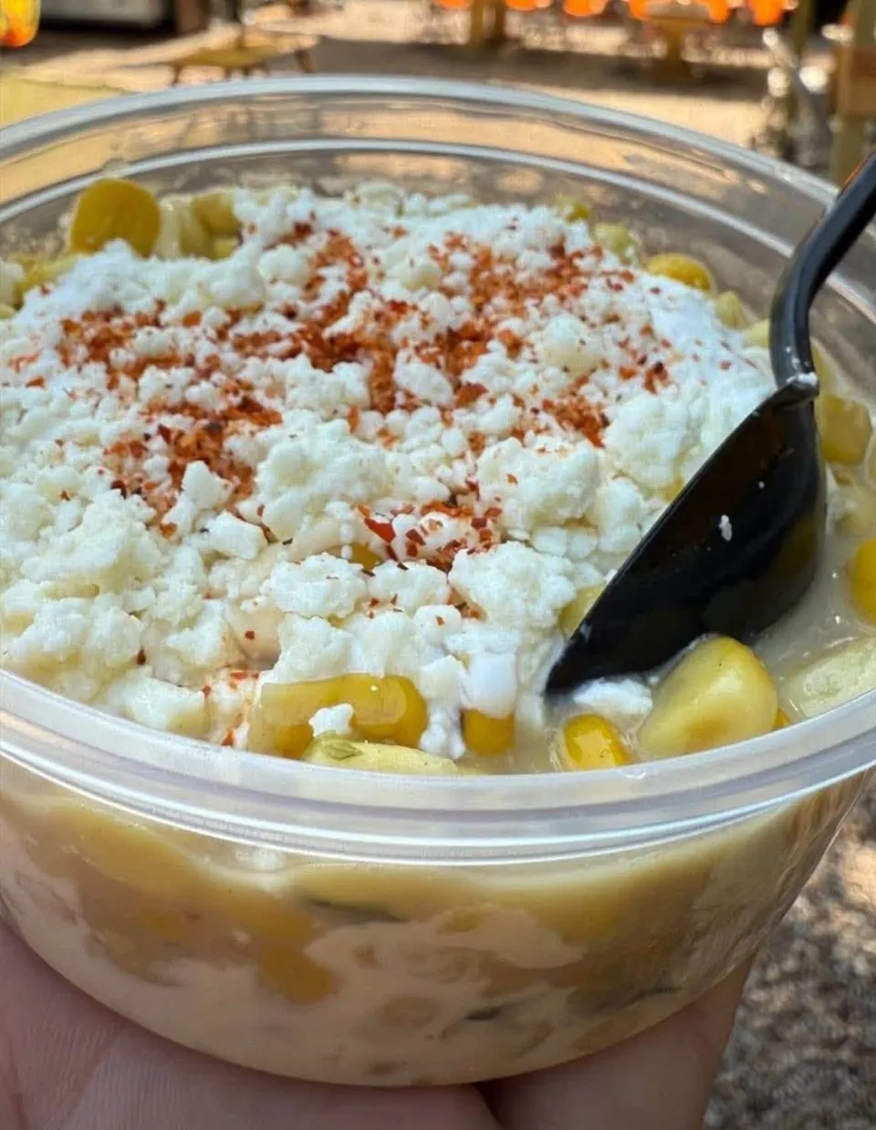 A cup of elote en vaso, creamy corn kernels topped with crumbled white cheese and red chile, with a black spoon.