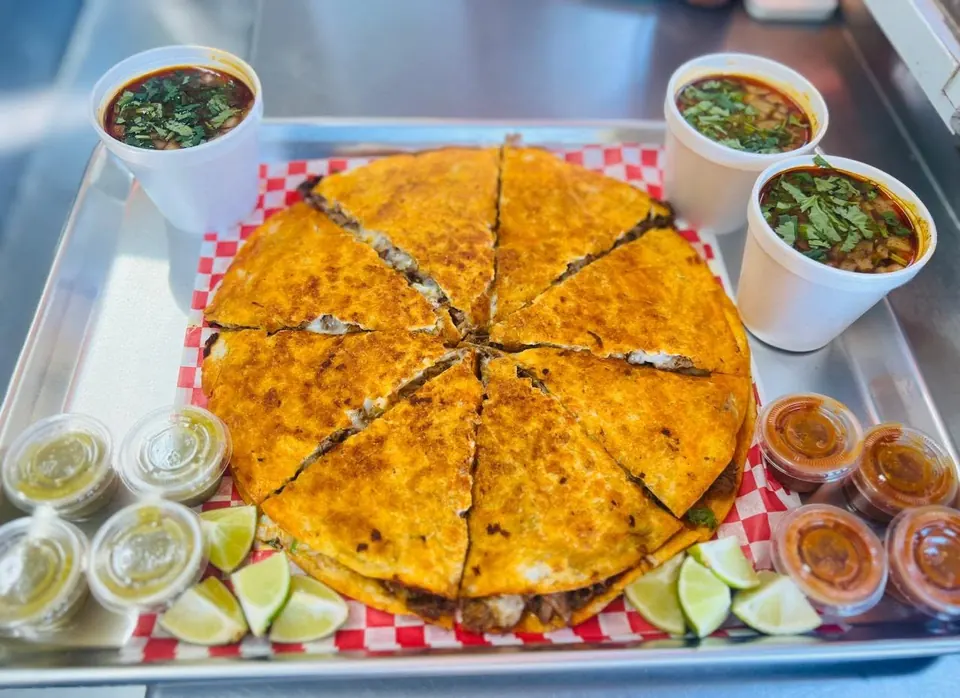 Pizza Birria cut into slices on a tray, with three cups of consommé, limes, and salsas.
