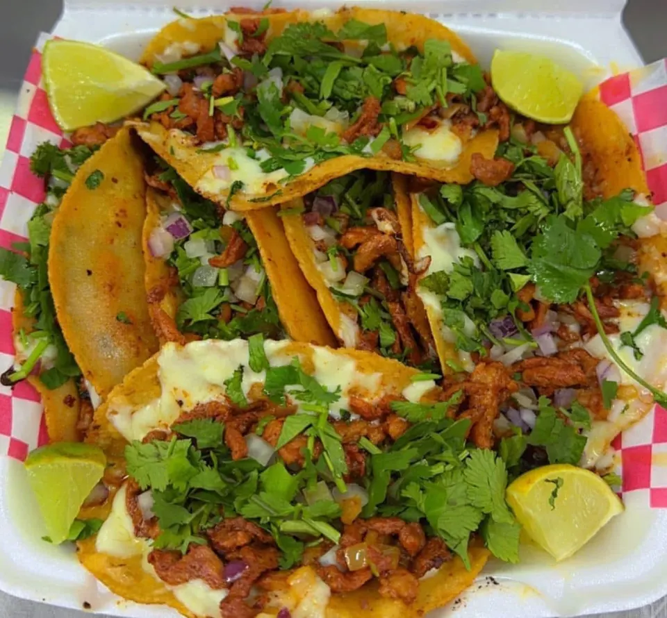 A plate of tacos with red-marinated pork, melted cheese, onions, cilantro, and lime wedges.