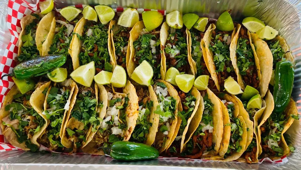 A foil tray packed with street tacos, lime wedges, and grilled jalapeños.