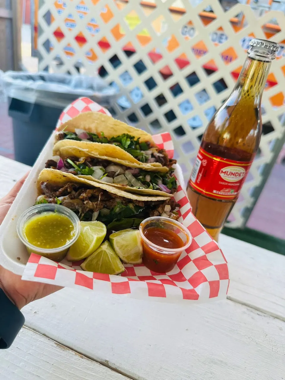 A hand holding a tray of tacos with salsas and lime next to a bottle of Mundet apple soda.