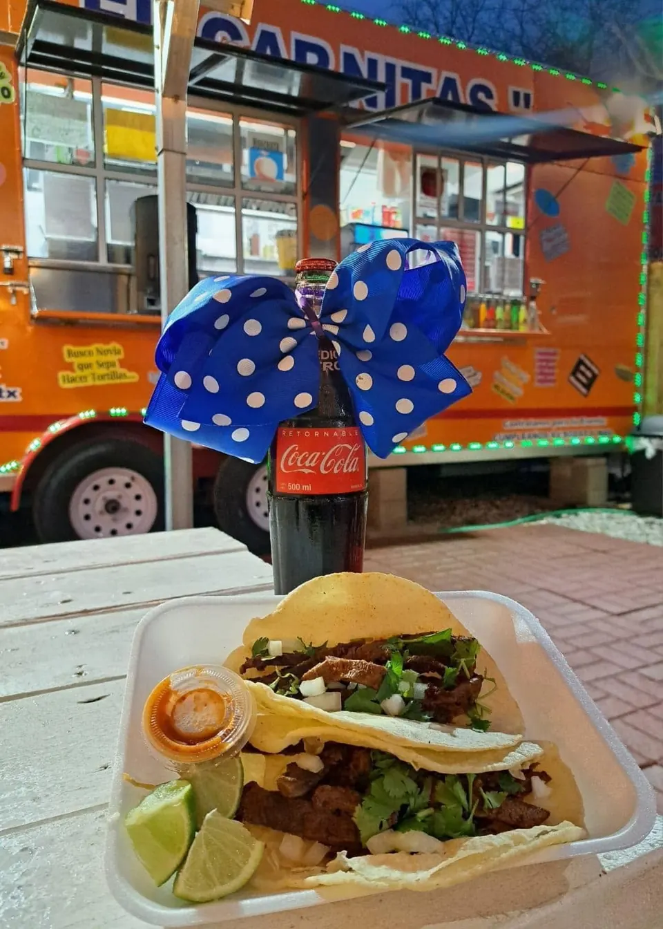 Two tacos topped with onions and cilantro, with lime and salsa, beside a glass bottle of Coca-Cola in front of the orange El Carnitas food truck.