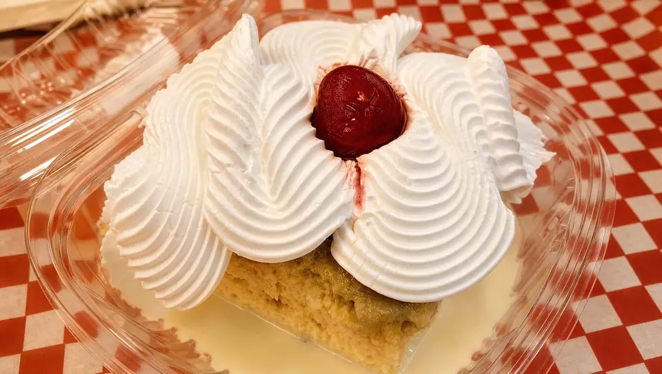 A slice of tres leches cake in a clear to-go container, piled with piped whipped cream and a cherry, sitting in milk on red checkered paper.